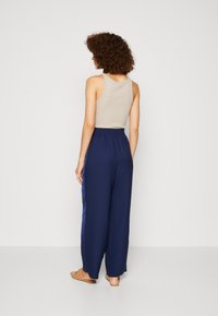 Pantalons larges bleu marine avec une taille élastique et une bande satinée lisse le long des côtés, associés à un débardeur beige et des mules.