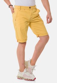 Gelbe Baumwollshorts mit umgeschlagenen Säumen, die mit roten Nähten verziert sind und Seitentaschen haben, kombiniert mit weißen und roten Sportschuhen.