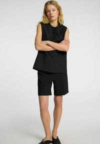 Černý bezramínkový top s knoflíkovým zapínáním a odpovídajícími tailored šortkami. Hladká tkanina, strukturovaný design, kapsy nahoře a ploché boty jako akcenty.