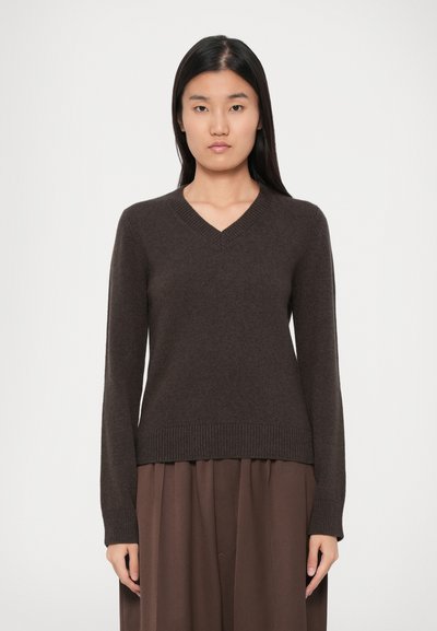 Femme avec des cheveux noirs raides, portant un pull V en marron foncé et un pantalon marron ample, se tenant devant un fond clair.