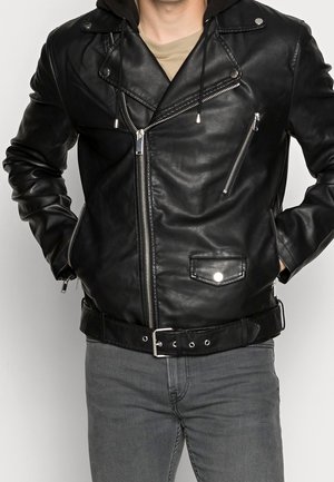 Sort læder bikerjakke med sølv lynlåse, trykknapkrave, bælte i taljen, båret over en tanfarvet skjorte og kombineret med grå jeans.