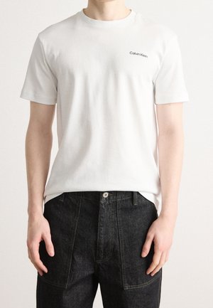Mand iført en enkel hvid kortærmet Calvin Klein t-shirt og sorte denim bukser mod en lys baggrund.