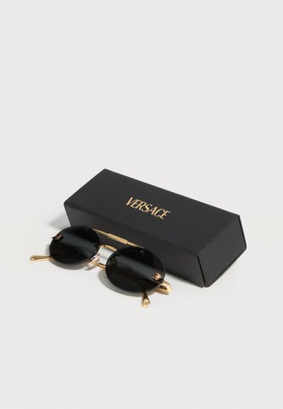 Στρογγυλά γυαλιά ηλίου με μαύρους φακούς και χρυσά πλαίσια, τοποθετημένα δίπλα σε μια μαύρη ορθογώνια θήκη με χρυσό λογότυπο Versace σε λευκή επιφάνεια.
