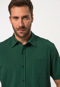 Camisa verde de manga corta en tela suave con cierre de botones en el frente, cuello clásico y un solo bolsillo en el pecho. Cuenta con botones negros.