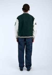 Giacca bomber verde e bianca con un colletto a coste, dotata di dettagli ricamati sulle maniche, abbinata a jeans a gamba larga blu scuro.