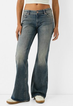 Femme portant un jean évasé bleu délavé, des baskets blanches et un débardeur court blanc sur un fond uni.