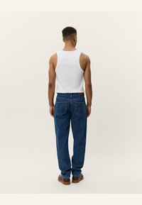Weißes ärmelloses Tanktop und blaue Jeans mit einer entspannten Passform, zwei Gesäßtaschen und brauem Schuhwerk. Schlichte, lässige Gestaltung.