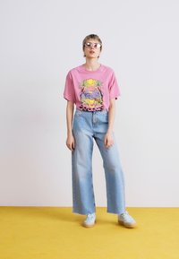 Pink graphic t-shirt with beach scene, wide-leg light denim jeans, and light blue sneakers, set against a yellow floor and white background.