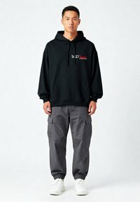Felpa nera con tasca frontale e testo "SWEET heart" in bianco e rosso, abbinata a pantaloni cargo grigi con tasche laterali e polsini elasticizzati. Sneakers bianche.