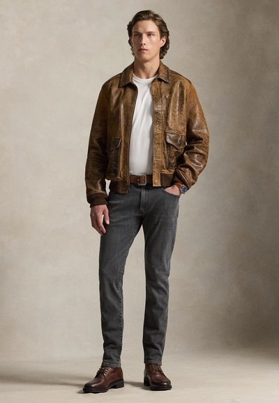 Νεαρός άνδρας που στέκεται φορώντας καφέ δερμάτινο μπουφάν, λευκό T-shirt, σκούρο γκρι τζιν, καφέ ζώνη, καφέ μπότες και ρολόι χειρός.