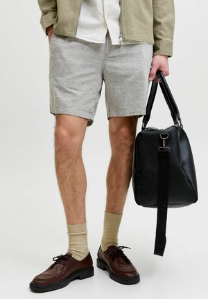 Mann trägt hellgraue Shorts, beige Socken, braune Lederschuhe und hält eine schwarze Lederreisetasche an den Griffen.