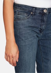 Nära bild av en person som bär blå denimjeans med synlig sömnad, frontknapp och bältesöglor, med vänster hand vilande vid sidan.
