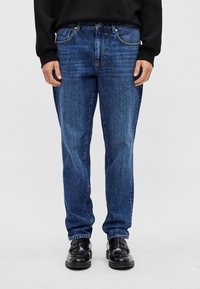 Vaqueros de mezclilla azul con un corte cómodo, que cuentan con cinco bolsillos, herrajes plateados y un desgastado sutil. Combinados con mocasines negros.