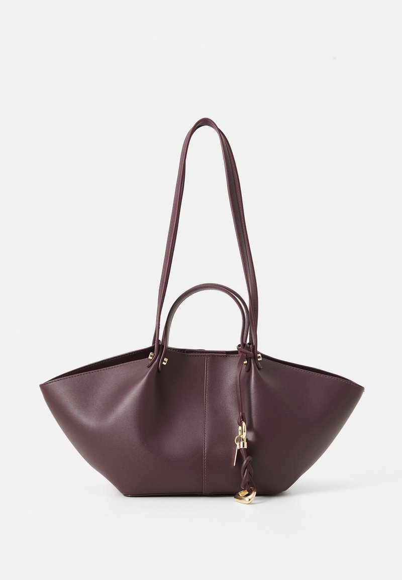 Sac fourre-tout en cuir bordeaux avec un design angulaire, deux longues sangles et une petite poignée. Présente des accents en métal doré et une fermeture à cordon.
