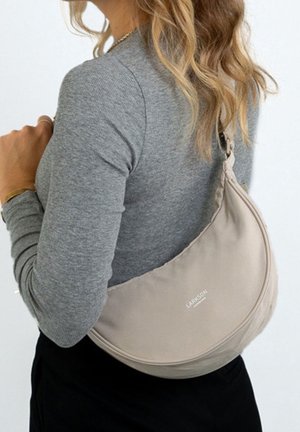 Pitkähiuksinen nainen, jolla on harmaa pitkähiha-paita ja joka kantaa beigeä Larksonin olkalaukkua selkänsä yli.