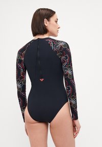 Maillot de bain à manches longues noir avec un motif fleuri sur les manches, fermeture éclair au dos et un petit emblème en forme de cœur dans le bas du dos.