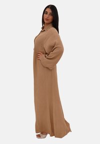 En kvinde står selvsikkert i en lang, beige kjole med vide ærmer, der viser et afslappet men stilfuldt look. Hun bærer hvide sandaler.