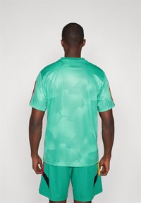 Camisola desportiva em malha verde com um padrão subtil, mangas curtas e detalhes vermelhos nos ombros. Acompanhada de calções verdes a condizer.