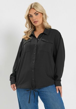 Blondine vrouw met een losse zwarte blouse met knopen en borstvoorraden en een blauwe spijkerbroek, staand tegen een egale lichte achtergrond.