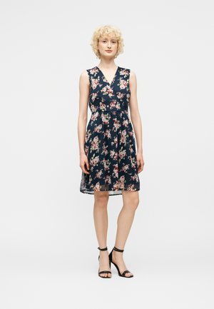 Kvinde med krøllet blondt hår iført en ærmeløs marineblå kjole med pink blomsterprint og sorte højhælede sko med ankelrem, stående foran en ensfarvet baggrund.