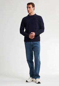 Marineblauer Strickpullover mit rundem Ausschnitt und gerippten Bündchen, kombiniert mit hellblauen Jeans und weißen Turnschuhen. Schlichtes Design, ohne Muster.