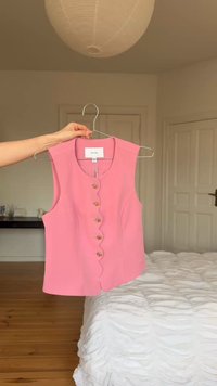 Main tenant un gilet sans manches rose avec une patte de boutonnage festonnée sur un cintre métallique dans une chambre neutre avec une literie blanche.