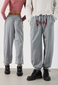 Grå joggebukser med elastiske mansjetter, med en lomme og et rødt grafisk design bak. Kombinert med svarte snøresko.