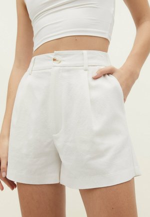 Femme portant un short blanc texturé taille haute avec un bouton et des poches latérales, associé à un débardeur court blanc.