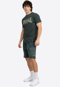 Mørk grøn T-shirt med "Lonsdale London" grafik, parret med matchende shorts med et løvelogo, hvide sneakers og ankelstrømper.