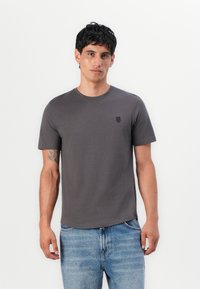 Graues T-Shirt mit Rundhalsausschnitt aus Baumwolle, kurzen Ärmeln und einem dezenten Logo auf der linken Brust, kombiniert mit hellblauen Jeans.