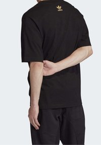 Svart bomulls-t-shirt med korta ärmar, med en guldfärgad Adidas-logotyp på baksidan och en avslappnad passform. Bärs med svarta byxor.