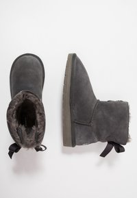 Šedé semišové kotníkové boty s měkkou kožešinovou podšívkou, plochými gumovými podešvemi a dekorativními černými stuhami na zadní straně.