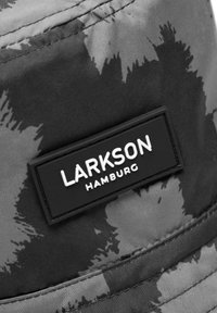 Černý a šedý kyblíkový klobouk s camouflage vzorem, který má obdélníkovou černou značku s bílým písmem a hladkým textilním povrchem.
