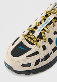 Τα αθλητικά παπούτσια Nike διαθέτουν σχέδιο από μπεζ και μαύρο δίχτυ με κίτρινα κορδόνια, μπλε λεπτομέρειες και μοναδική υφή στη σόλα.