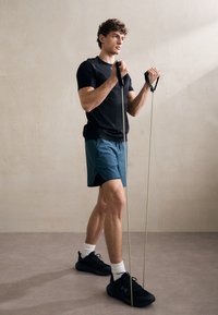 Črn športni majica in modri šort. Moški drži elastične trakove z črnimi ročaji. Obut v črne čevlje in bele nogavice. Enobarven ozadje.