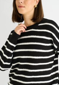 Black and white striped sweater with a relaxed fit, textured knit pattern, ribbed cuffs, and a wide neckline. Gold hoop earrings complement the look.