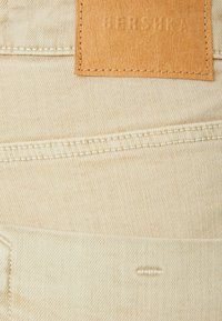 Beige denimtyg med en synlig läderetikett, sydd med kontrasterande vit tråd. Har bakfickor och en subtil texturdetalj.