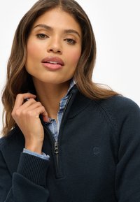 Marine-blauwe zip-up trui van breimateriaal, met een hoge kraag, geribbelde manchetten en een klein logo op de borst. Gelaagd over een blauw geruit overhemd.
