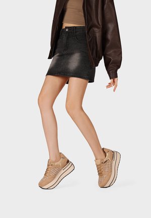 Donna con sneakers beige con piattaforma, minigonna nera di denim, top corto marrone e giacca di pelle marrone scuro, in piedi su una gamba.