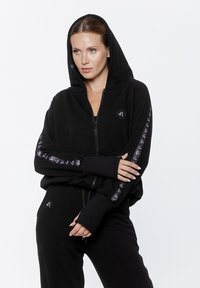Czarna bluza z kapturem na zamek błyskawiczny, z podszewką w kapturze, z długimi rękawami z akcentami wzoru logo i otworami na kciuki, wykonana z miękkiego, teksturowanego materiału.