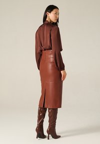 Blusa di raso marrone con maniche lunghe, abbinata a una gonna a matita in pelle marrone a vita alta con uno spacco posteriore e stivali marroni con tacco al ginocchio.