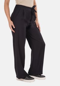 Pantaloni neri a gamba larga realizzati in un tessuto leggero, con pieghe e vita legata. Il modello li abbina a sneakers nere.