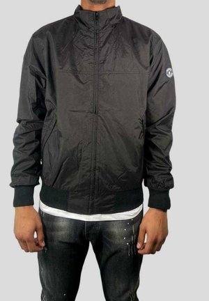 Man die een zwarte bomberjack draagt met een ritssluiting, zakken, geribbelde manchetten en zoom, over een wit overhemd en donkere spijkerbroek.