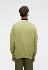 Prevelik kroj sweatshirt z okroglim izrezom v svetlo olivno zeleni barvi, z rebrastimi manšeti in obrobo, z črnimi ovratnikom, ki je položen pod njim.