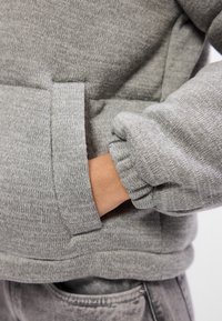 Person iført grå strikket sweatshirt med hånden i forreste lomme og elastiske manchetter mod en lys baggrund.