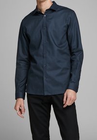 Man i en välsittande mörkblå skjorta med knappar och långa ärmar samt svarta byxor mot en neutral bakgrund.