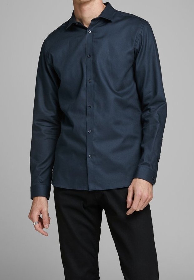 Man i en välsittande mörkblå skjorta med knappar och långa ärmar samt svarta byxor mot en neutral bakgrund.
