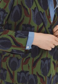 Blazer tricoté avec des motifs de tulipes bleu foncé sur un fond vert, doté d'un col cranté et de poignets de chemise bleu clair.