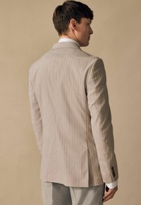 Light beige pinstriped blazer made of fabric with a smooth texture. Features a single vent, tailored fit, and dark button accents.