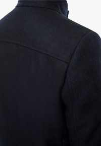 Black wool coat featuring a textured finish, high collar, and cleanly sewn seams, emphasizing a tailored silhouette and structured design.